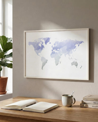 A framed world map art print with watercolor-style continents in shades of lavender and blue on a white background, hanging on a beige wall above a wooden desk with an open book, mug, and stacked books.