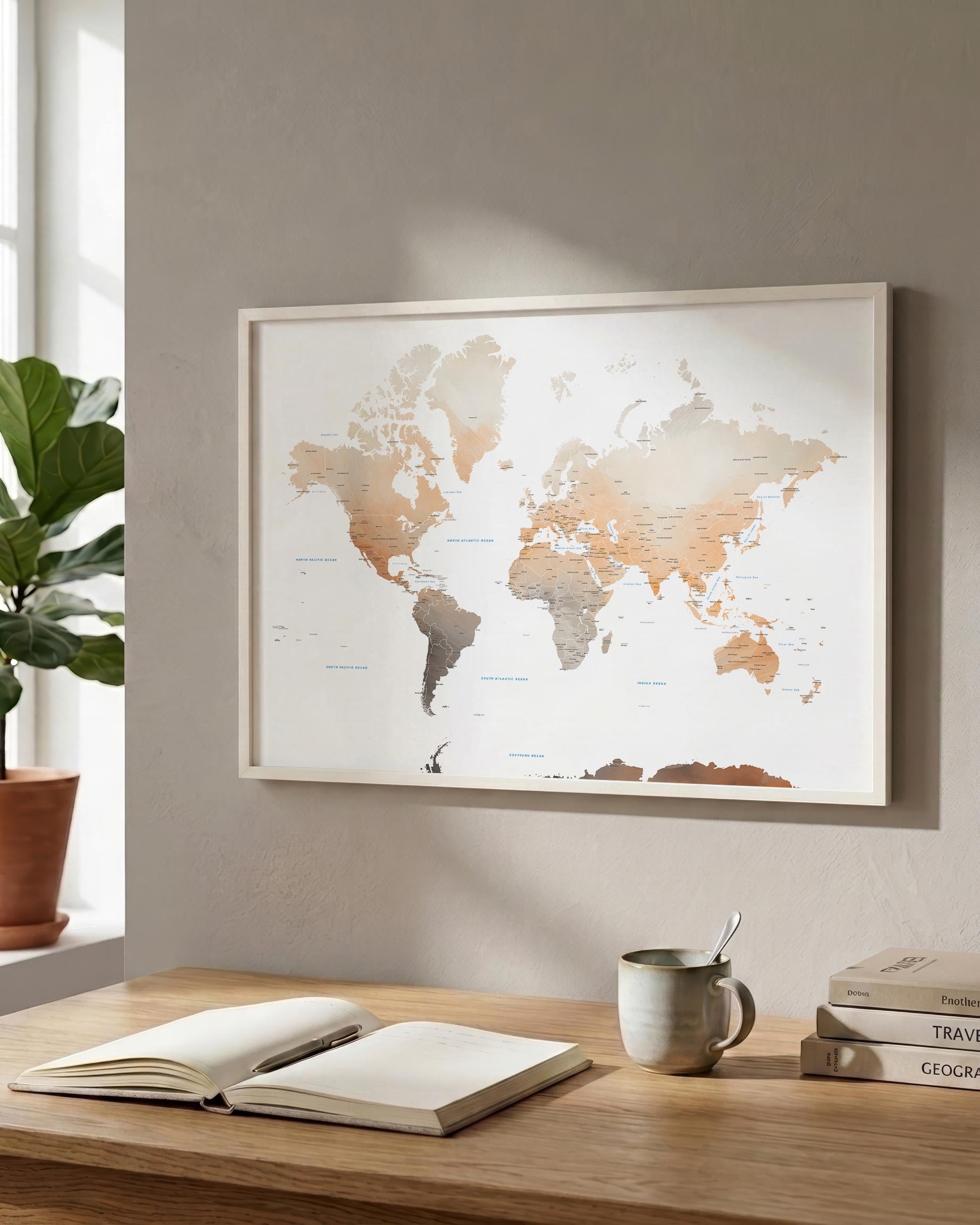 A framed world map print with muted beige, brown, and gray landmasses hanging on a light gray wall above a wooden desk with an open book, mug, and stacked books.