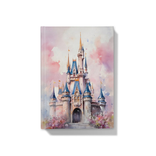 A person holds a notebook featuring a pastel watercolor illustration of a castle, resting on a cozy beige knit blanket on a wooden table with a cup of tea nearby.