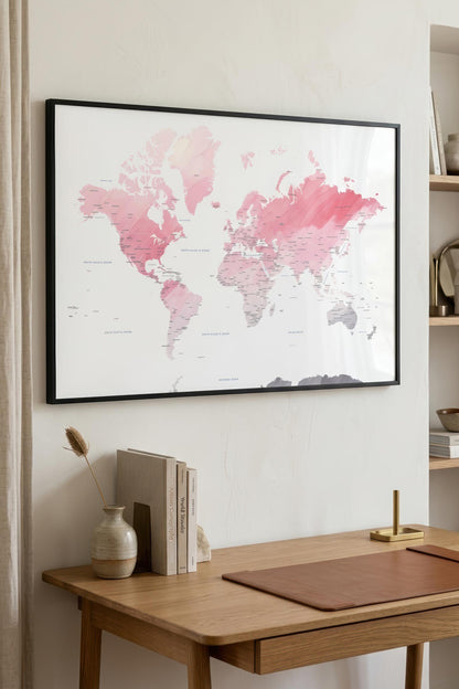 A framed world map print with pink watercolor continents and gray labels, displayed on a light beige wall above a wooden desk with books and a vase.