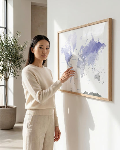 A woman in a cream knit sweater and matching pants stands beside a large framed world map hanging on a white wall in a bright, modern room.