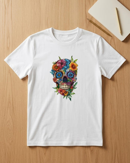 A white short-sleeve crewneck t-shirt featuring a colorful sugar skull graphic with flowers, laid flat on a light wood background next to a notebook and pencil.
