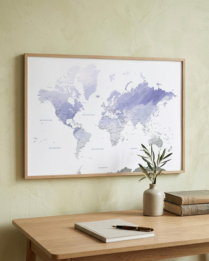 A framed world map print with a light wood frame hangs on a textured cream wall above a wooden desk with a notebook, pen, and a small vase with green stems.