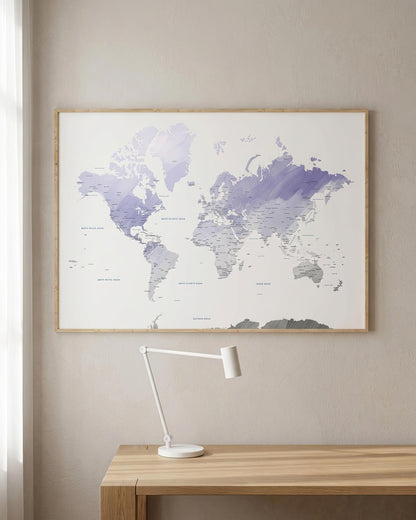 A framed world map poster with a watercolor-style purple and gray landmass on a white background, hanging on a beige wall above a wooden desk with a white desk lamp.