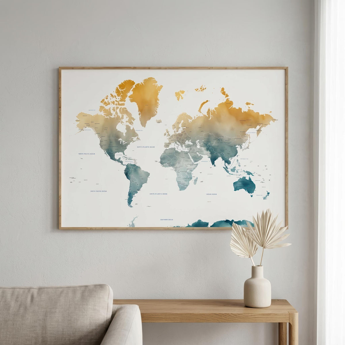 A framed world map wall art print hangs on a light gray wall above a wooden console table with a vase of dried leaves. The map features watercolor-style continents in gold, orange, and teal on a white background, enclosed in a thin natural wood frame.