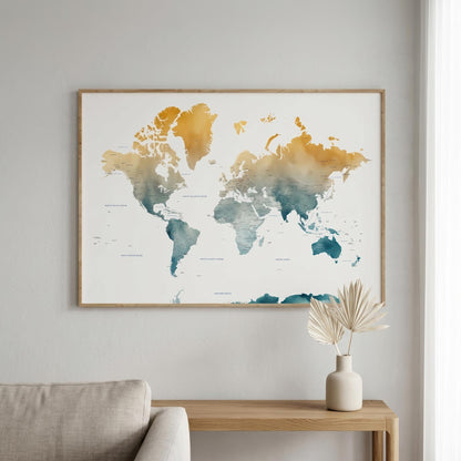A framed world map wall art print hangs on a light gray wall above a wooden console table with a vase of dried leaves. The map features watercolor-style continents in gold, orange, and teal on a white background, enclosed in a thin natural wood frame.