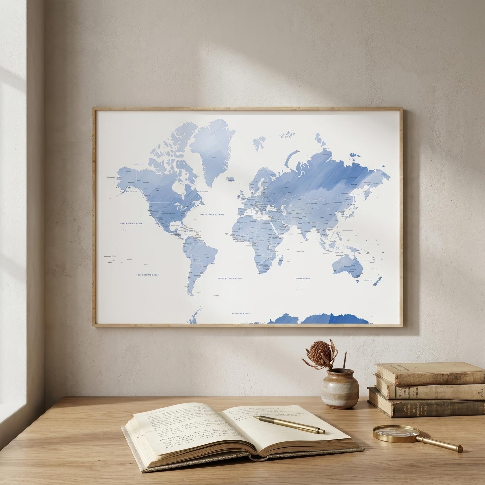 A framed world map poster with blue watercolor-style landmasses on a white background, hanging on a beige wall above a wooden desk with an open book, magnifying glass, and small vase.