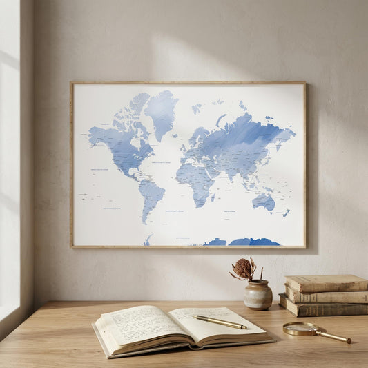 A framed world map poster with blue watercolor-style landmasses on a white background, hanging on a beige wall above a wooden desk with an open book, magnifying glass, and small vase.