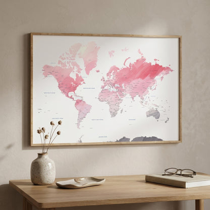 A framed world map wall art print with pink and coral watercolor continents on a white background, hanging on a beige wall above a wooden table with a vase, dish, and book.