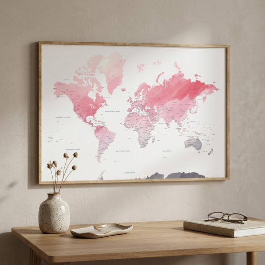 A framed world map wall art print with pink and coral watercolor continents on a white background, hanging on a beige wall above a wooden table with a vase, dish, and book.