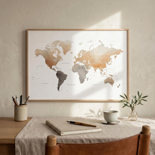 A framed world map print hangs on a textured beige wall above a styled desk with a notebook, cup, and small plant. The map features muted continents in warm beige, brown, and gray tones on a white background, enclosed in a light wood frame.