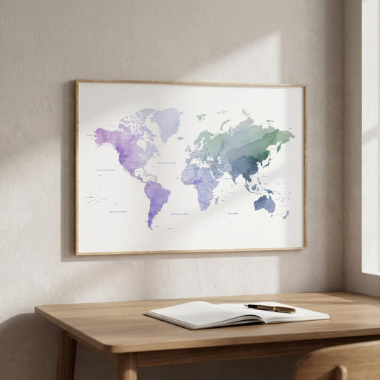 A framed world map art print with watercolor-style continents in purple, blue, and green tones, hanging on a light beige wall above a wooden desk with an open notebook and pen.