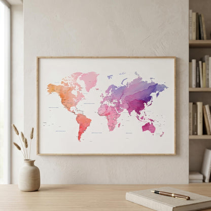 A framed world map poster with a watercolor design in pink, orange, and purple tones, displayed on a light beige wall above a wooden desk with a vase and notebook.
