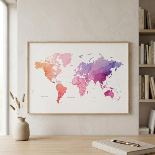 A framed world map poster with a watercolor design in pink, orange, and purple tones, displayed on a light beige wall above a wooden desk with a vase and notebook.