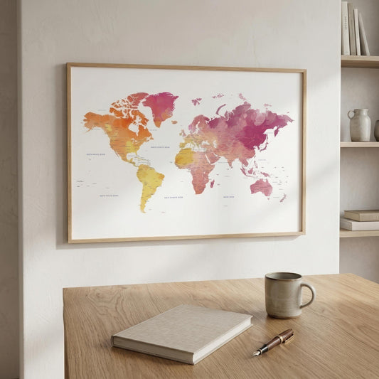 A framed world map watercolor print with warm multicolor continents hangs on a light beige wall above a wooden desk with a notebook, pen, and mug.