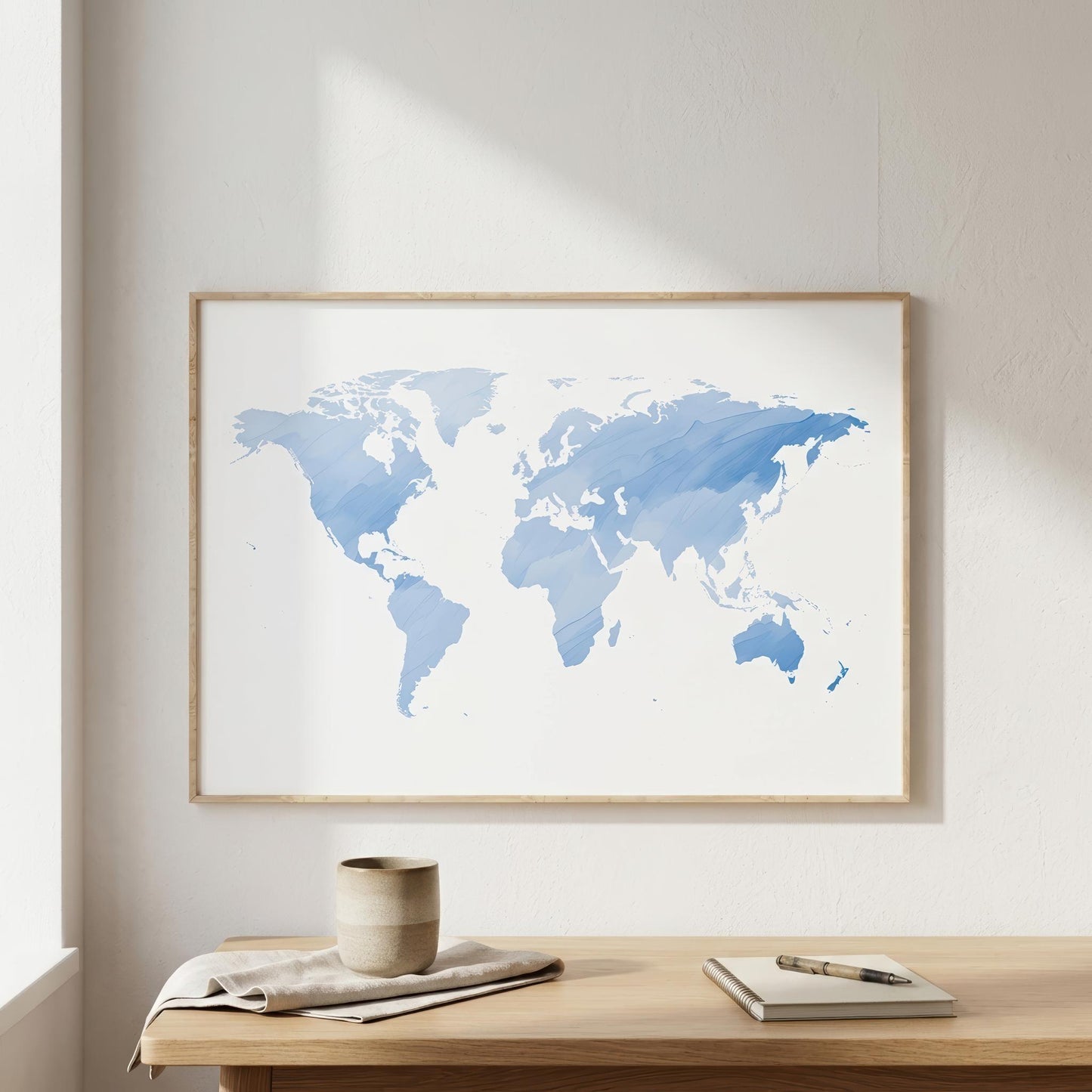 A framed world map art print in watercolor blue tones, hanging on a light beige wall above a wooden table with a mug, notebook, and pen.