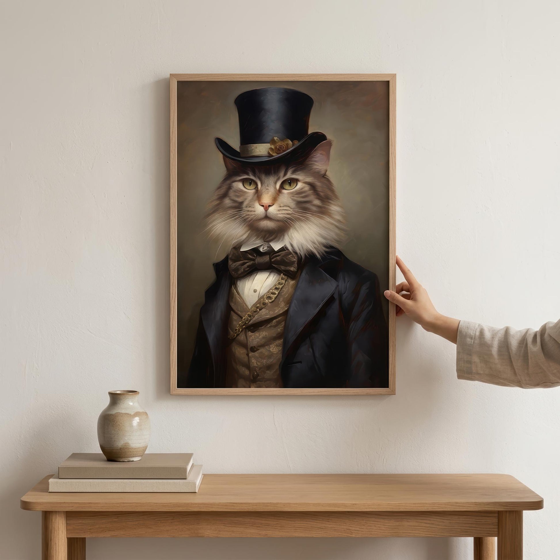 A framed vertical portrait print of a cat dressed in formal Victorian attire hangs on a light beige wall above a wooden console table. A human hand is touching the right side of the frame, and a small vase and books sit on the table.