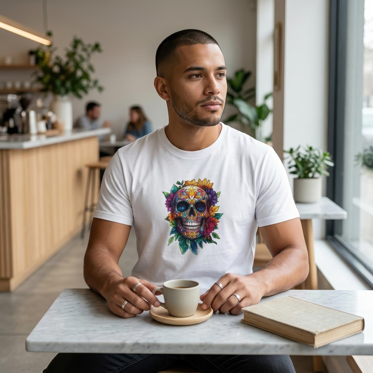 A man sitting at a cafe table wearing a white t-shirt with a colorful sugar skull graphic, holding a cup and saucer next to a book.