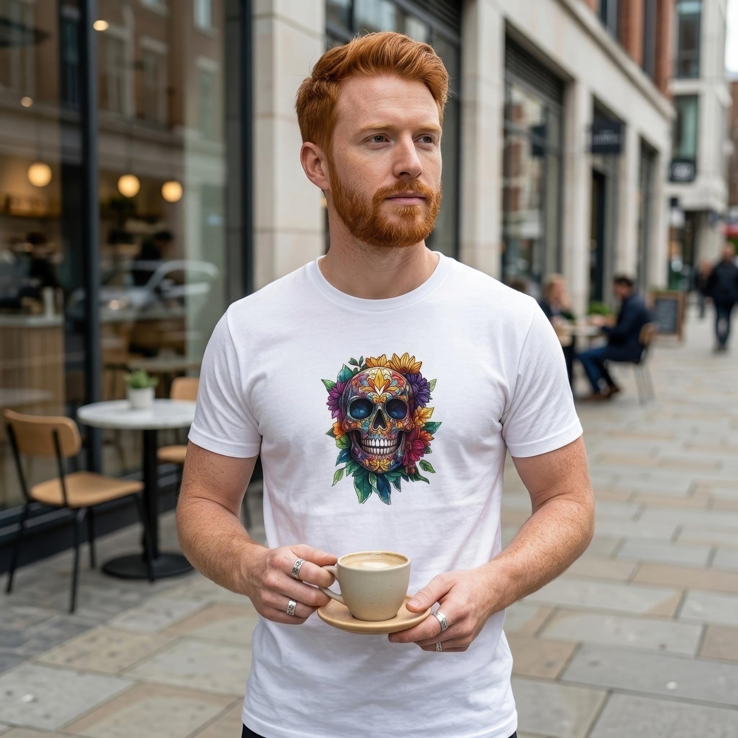 A man with red hair and a beard wearing a white graphic t-shirt with a colorful sugar skull design, holding a cup of coffee on a sidewalk in front of a cafe.