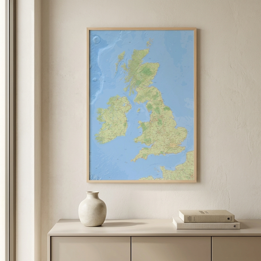 A framed topographic map of the United Kingdom and Ireland, featuring light blue oceans and green landmasses, hanging on a textured beige wall above a light wood console table with a ceramic vase and books.