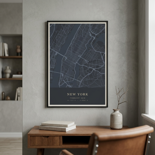 A framed New York City street map poster hangs on a textured gray wall above a wooden desk with books and a ceramic vase. The poster features a dark slate blue background with white map lines and gold text reading 'NEW YORK' and 'FEBRUARY 2025'.