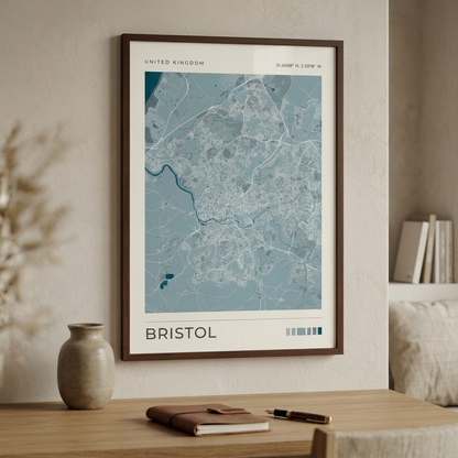 A framed map print of Bristol, United Kingdom, displayed on a beige wall above a wooden table with a notebook, pen, and vase.
