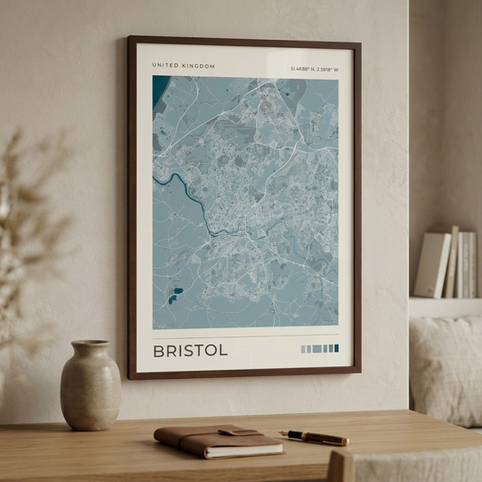A framed map print of Bristol, United Kingdom, displayed on a beige wall above a wooden table with a notebook, pen, and vase.