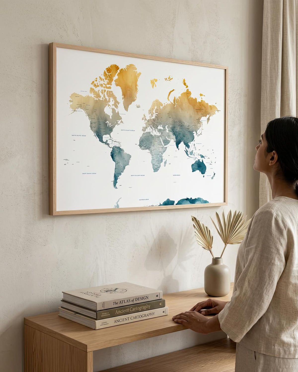 A woman in a neutral-toned outfit stands beside a light wood console table, looking at a framed world map wall art print hanging on a textured beige wall. The map features watercolor-style continents in gold, teal, and blue on a white background, with a light wood frame.