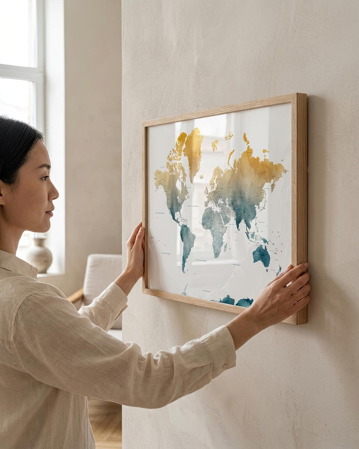 A woman in a beige linen shirt hangs a framed world map print on a textured beige wall. The map features watercolor continents in gold and teal on a white background, enclosed in a light wood frame.