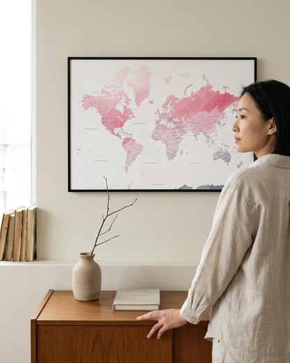 A woman in a light beige linen shirt stands beside a wooden cabinet, looking at a framed world map wall art print with pink watercolor continents. The room has a warm neutral tone with a vase and books on the cabinet.