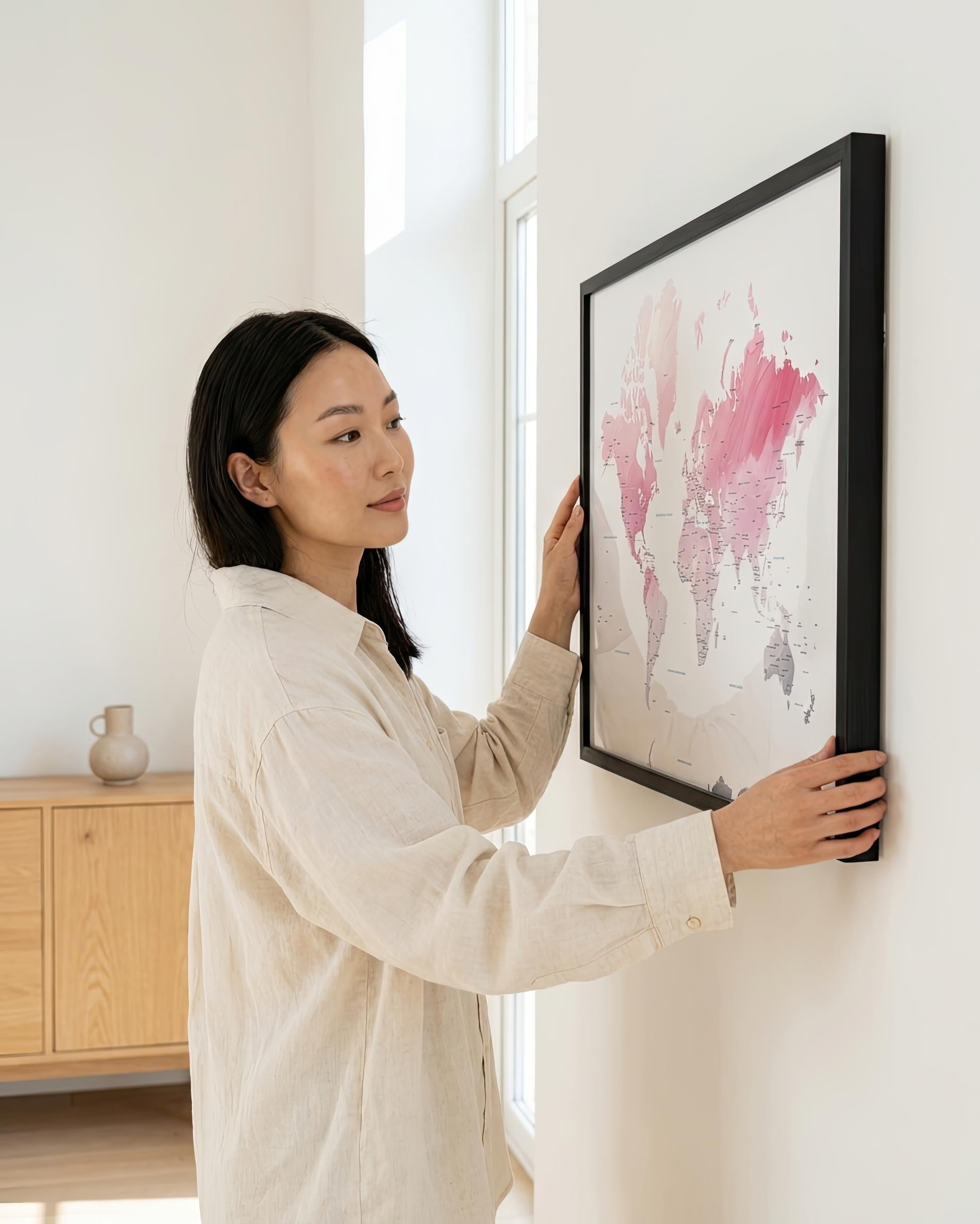 A woman in a light beige linen shirt hangs a large black-framed abstract world map print on a white wall in a bright, modern room.