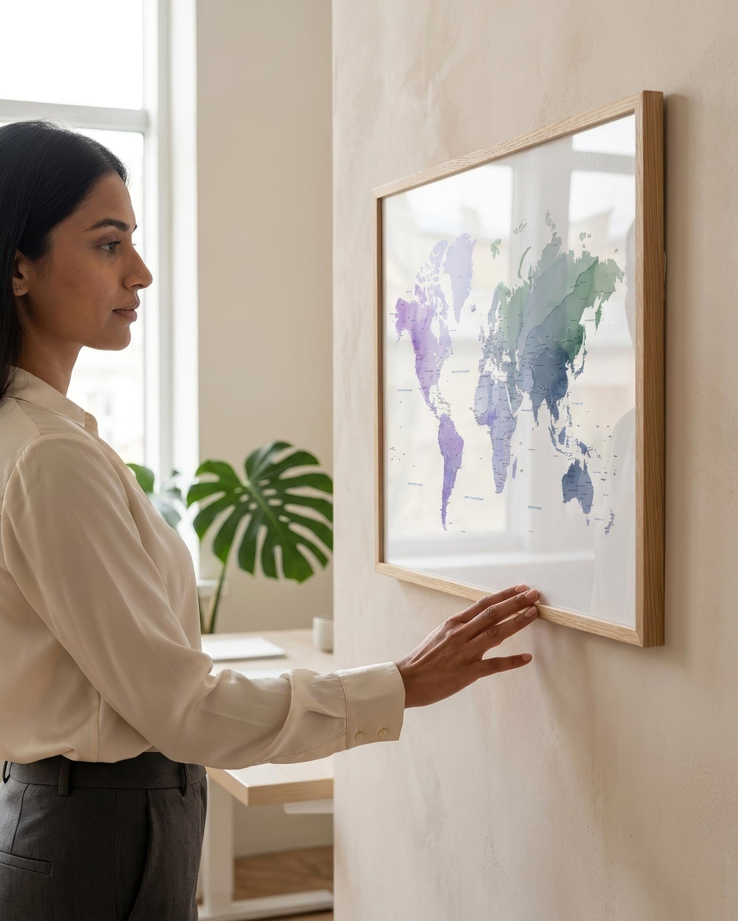 A woman in a cream blouse and dark trousers stands beside a framed world map hanging on a light beige wall. The map has a light wood frame and a white background with watercolor-style continents in purple, blue, and green.