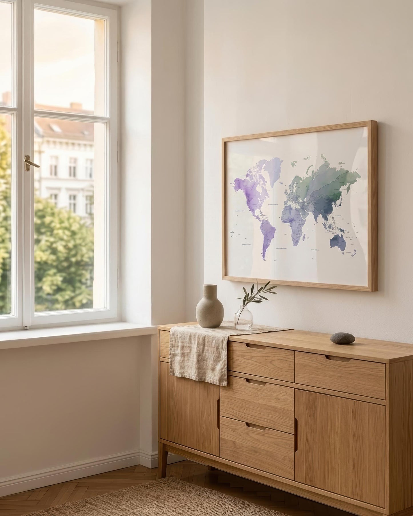 A framed world map print with watercolor-style continents in purple, blue, and green tones hangs on a light beige wall above a light wood sideboard in a bright room with a window.