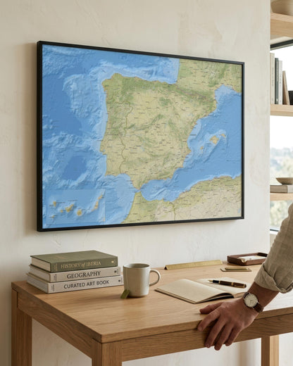 A framed wall map of Spain and surrounding sea hangs above a wooden desk in a bright room. A person's arm and hand are visible at the right edge, resting near the desk.