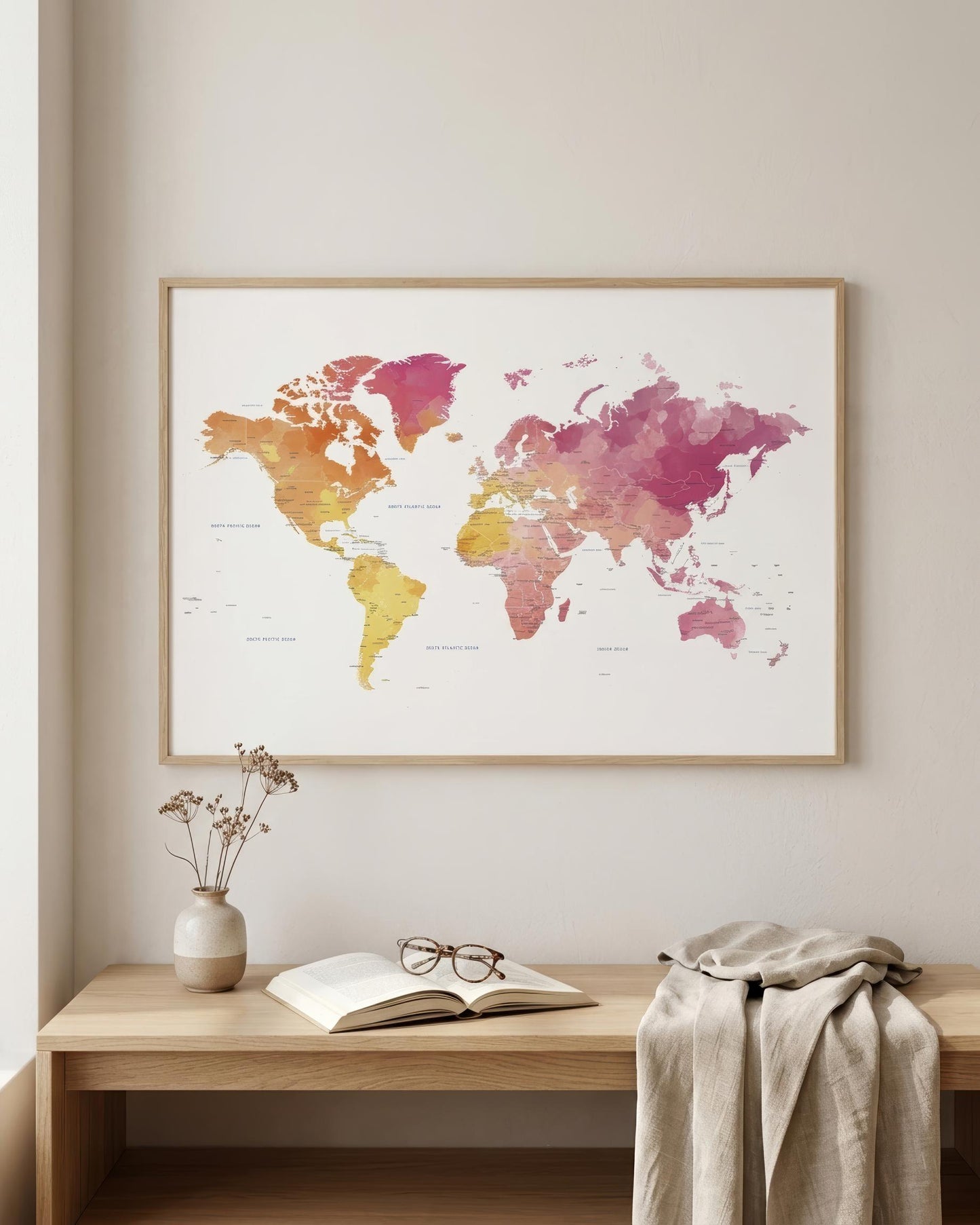 A framed world map watercolor print in pink, orange, and yellow tones, displayed on a light beige wall above a wooden console table with an open book, glasses, and a draped beige throw.