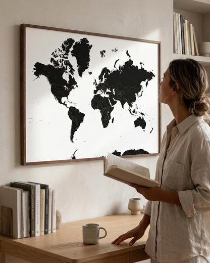 A woman in a light beige linen shirt stands beside a wooden desk, looking at a large framed world map wall art print. The map features black continents on a white background with a natural wood frame.