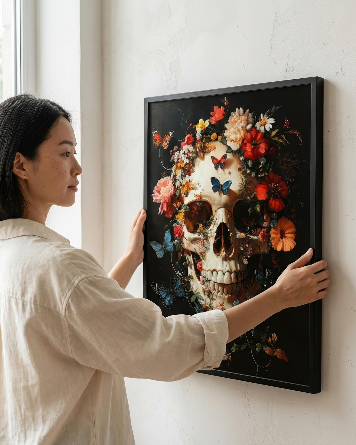 A woman in a beige shirt hangs a framed wall art print on a white wall. The artwork features a decorative skull surrounded by colorful flowers and butterflies on a dark background.