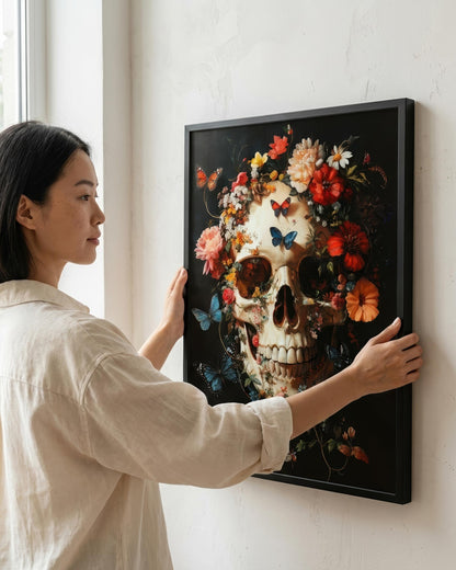 A woman in a beige shirt hangs a framed wall art print on a white wall. The artwork features a decorative skull surrounded by colorful flowers and butterflies on a dark background.