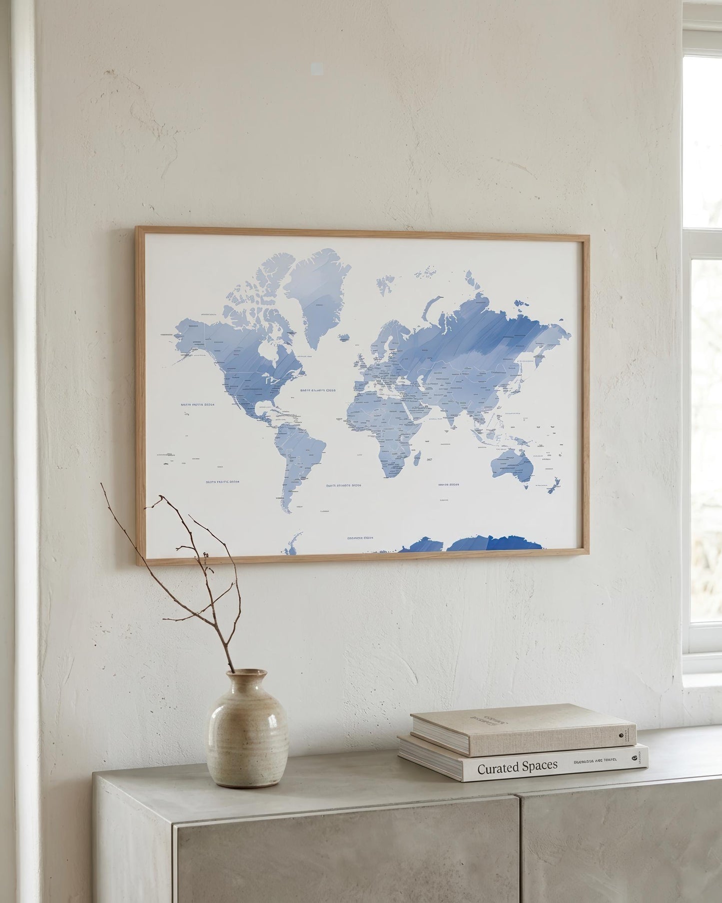 A framed world map print with blue watercolor continents on a white background, hanging on a light beige wall above a console table with a vase and books.