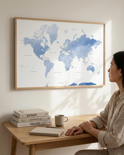 A woman sits at a wooden desk looking at a large framed world map hanging on the wall. The map features blue watercolor-style continents on a white background with a light wood frame.