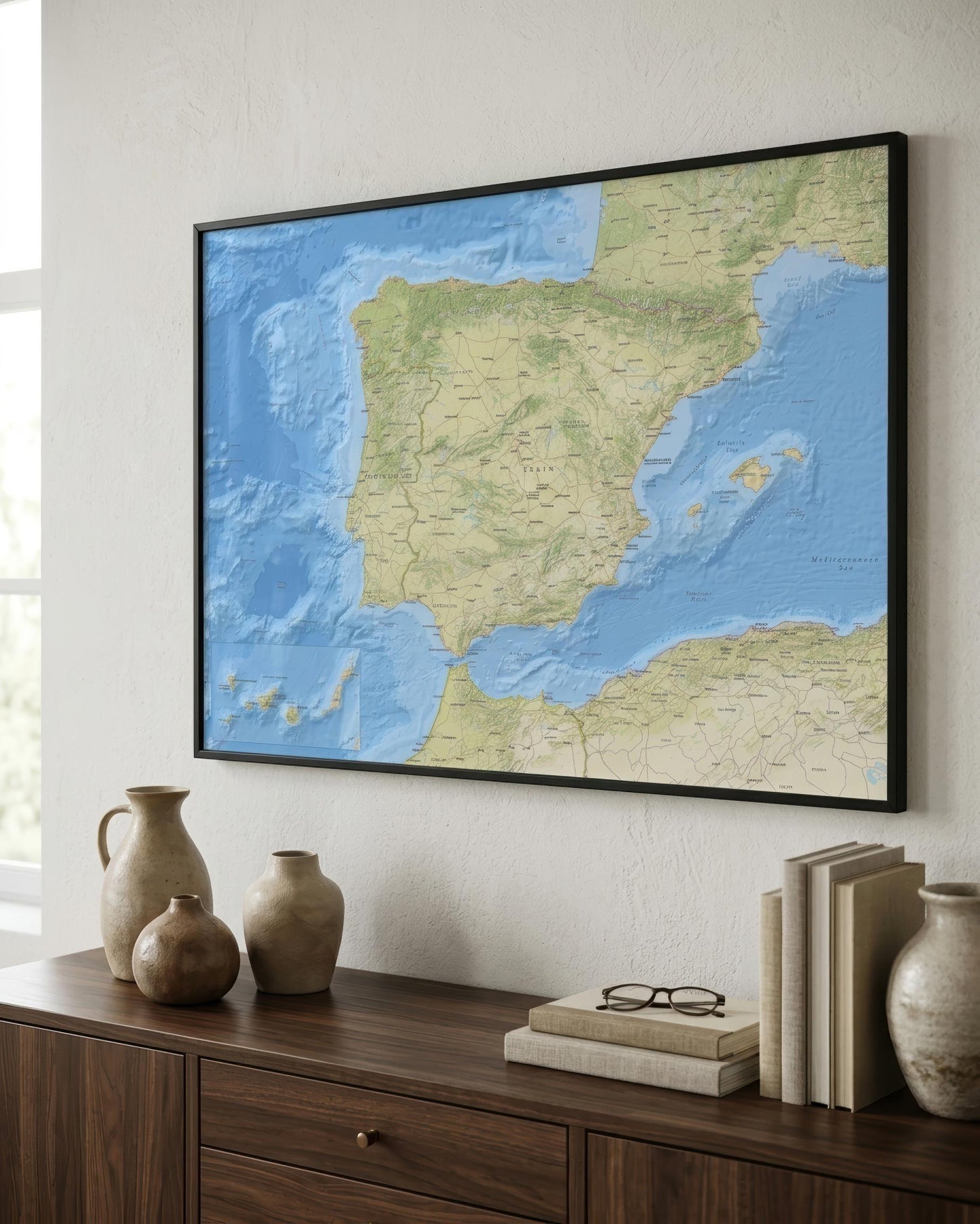 A framed 3D relief map of Spain and Portugal hanging on a light-colored wall above a wooden console table decorated with ceramic vases and books.