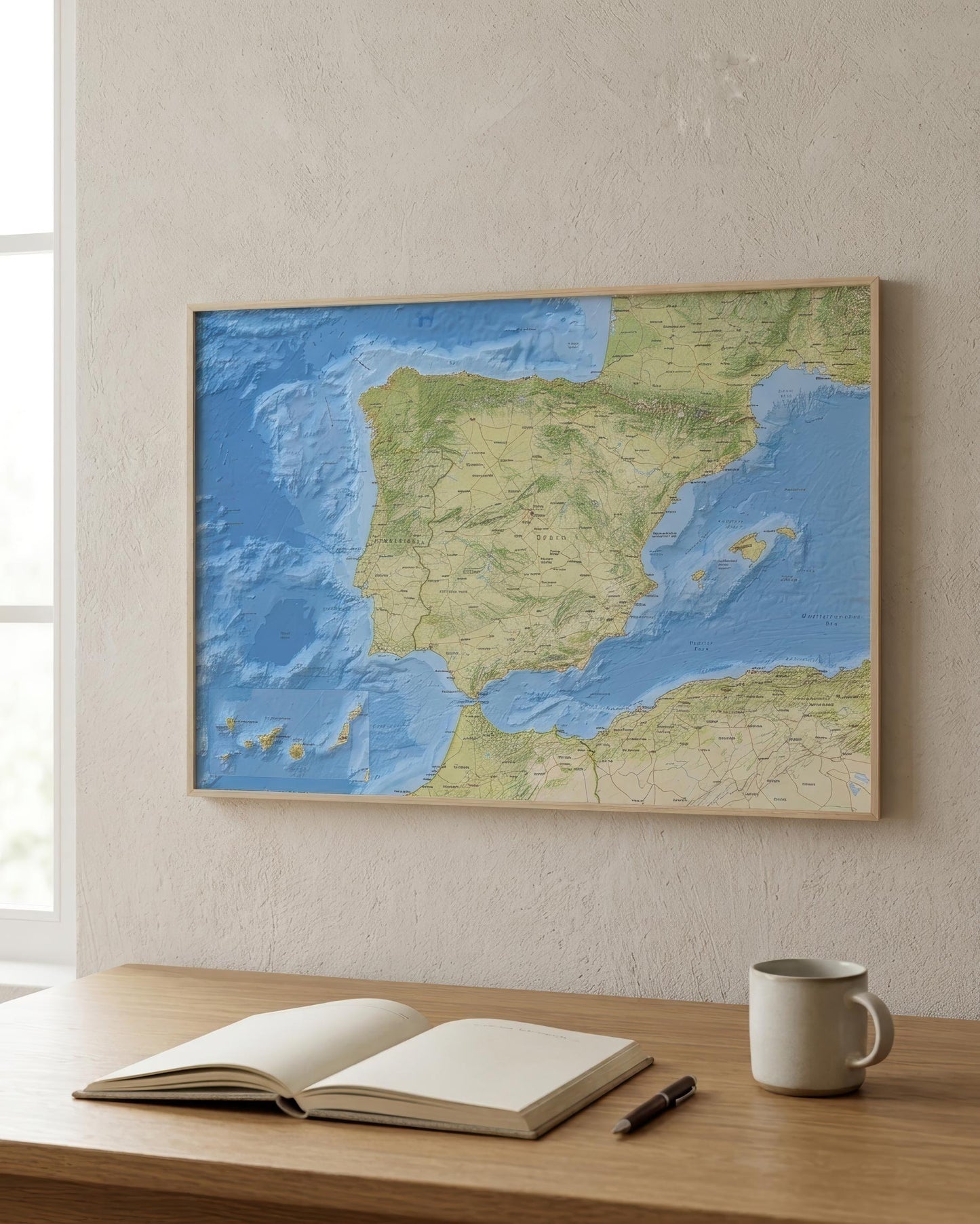 A framed topographic map of Spain and Portugal hanging on a textured beige wall above a wooden desk with an open notebook, a pen, and a white mug.