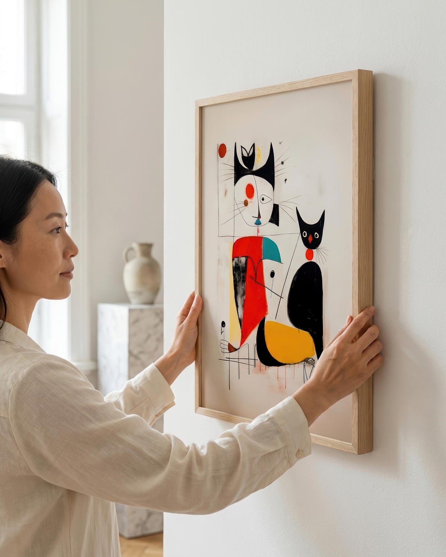 A woman in a cream linen shirt hangs a framed abstract art print featuring stylized black cats with red, yellow, and blue shapes on a white wall.