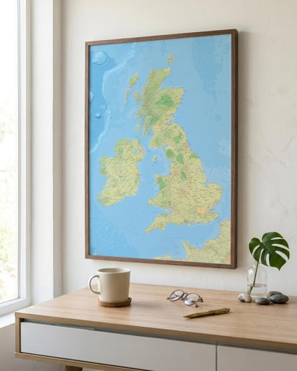 A framed map of the United Kingdom and Ireland, featuring light blue oceans and green landmasses, hanging on a beige wall above a wooden desk with a mug, glasses, and a small plant.