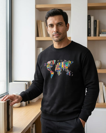 A man wearing a black crewneck sweatshirt with a colorful world map graphic made of various national flags, standing in a bright room with bookshelves in the background.