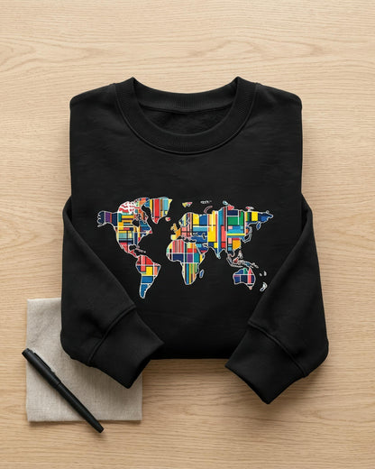 A black crewneck sweatshirt laid flat on a light wood surface, featuring a world map graphic filled with colorful national flags. A small beige napkin with a black pen sits to the lower left of the sweatshirt.