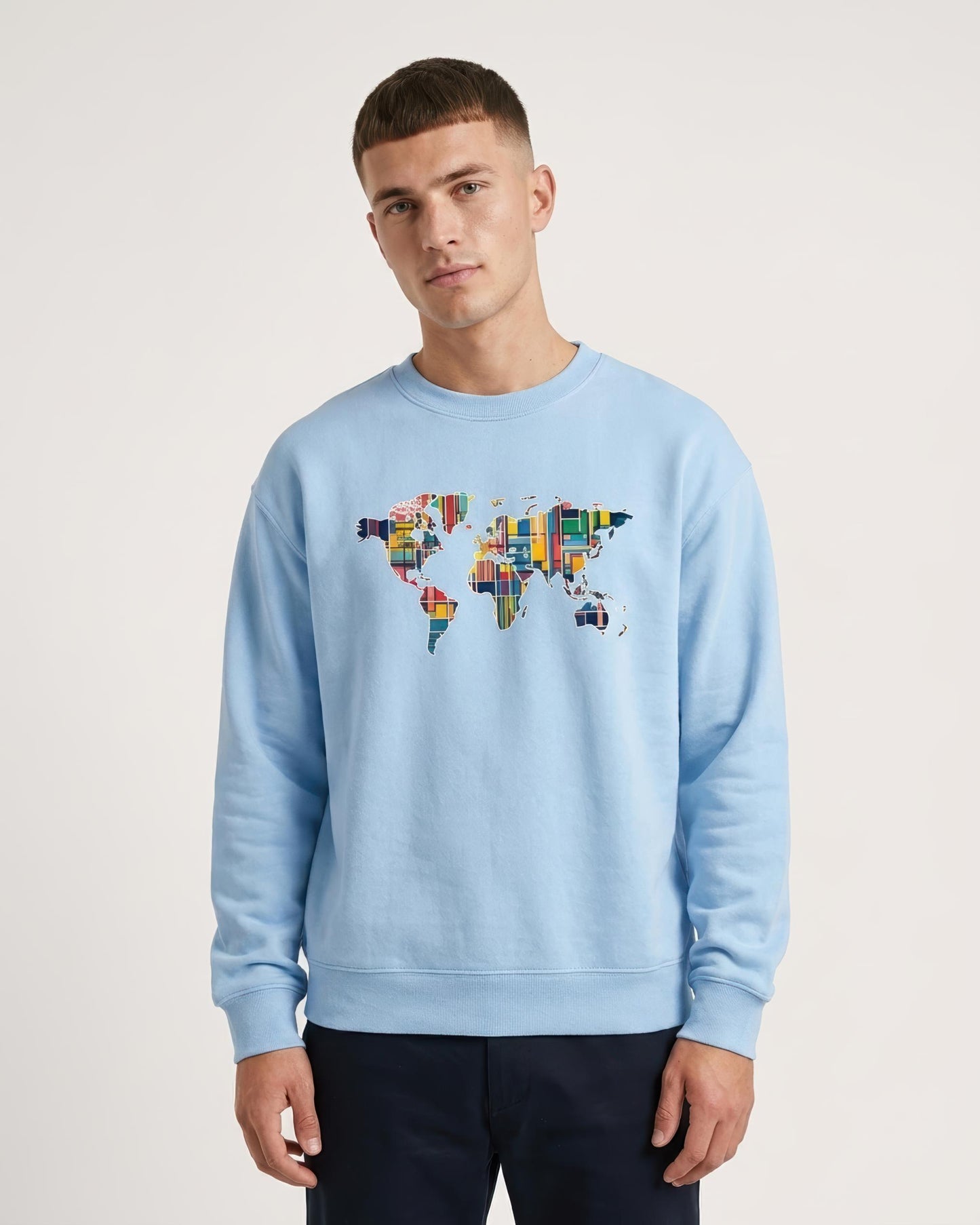 A man wearing a light blue crewneck sweatshirt with a colorful world map graphic on the chest, paired with dark navy pants, against a plain light gray background.