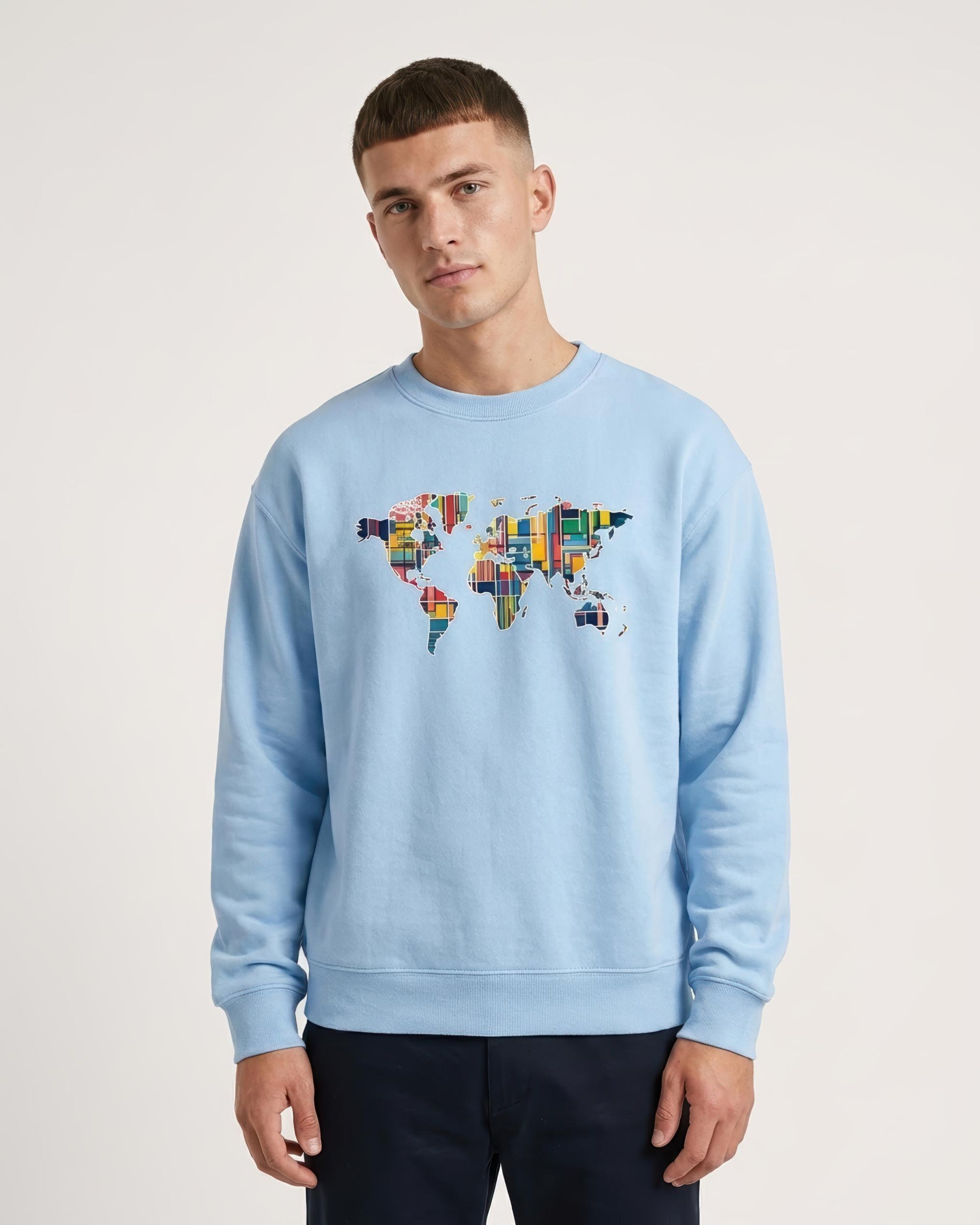 A man wearing a light blue crewneck sweatshirt with a colorful world map graphic on the chest, paired with dark navy pants, against a plain light gray background.