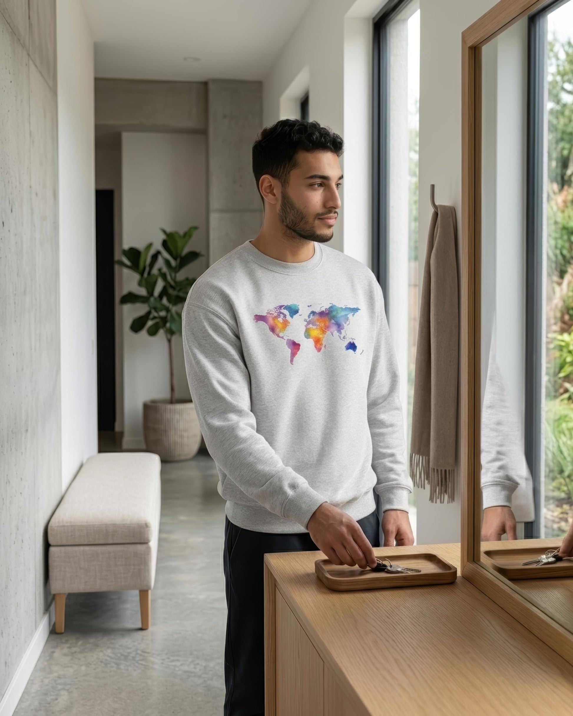 A man wearing a light gray crewneck sweatshirt with a colorful watercolor world map graphic on the chest, standing in a modern minimalist hallway.