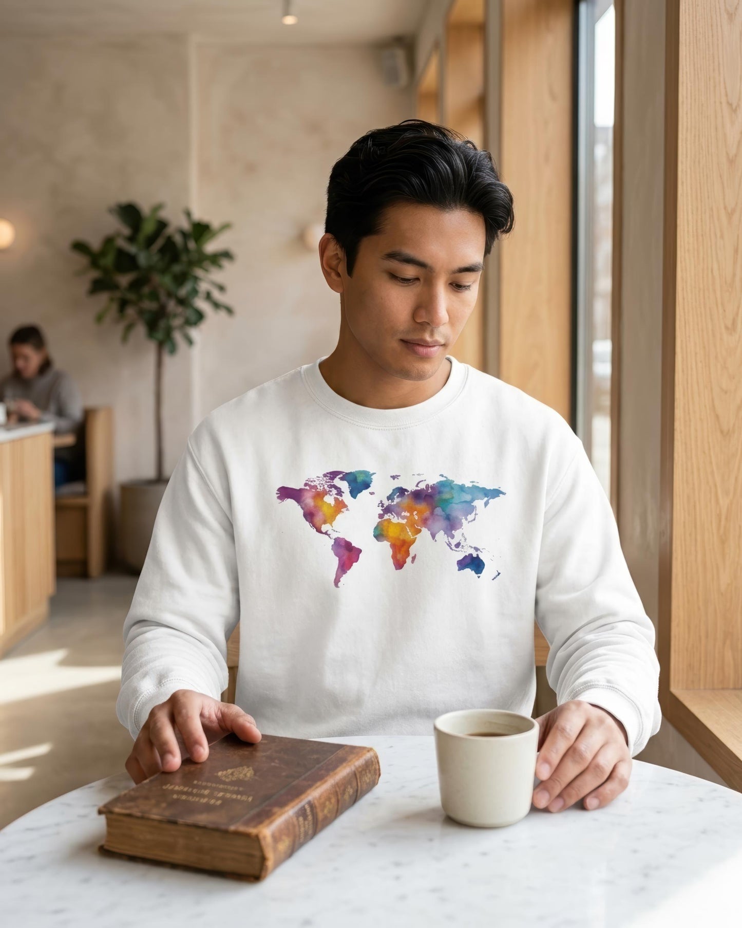 A man wearing a white crewneck sweatshirt with a colorful watercolor world map graphic on the chest, sitting at a table with a coffee mug and an old book in a cafe setting.